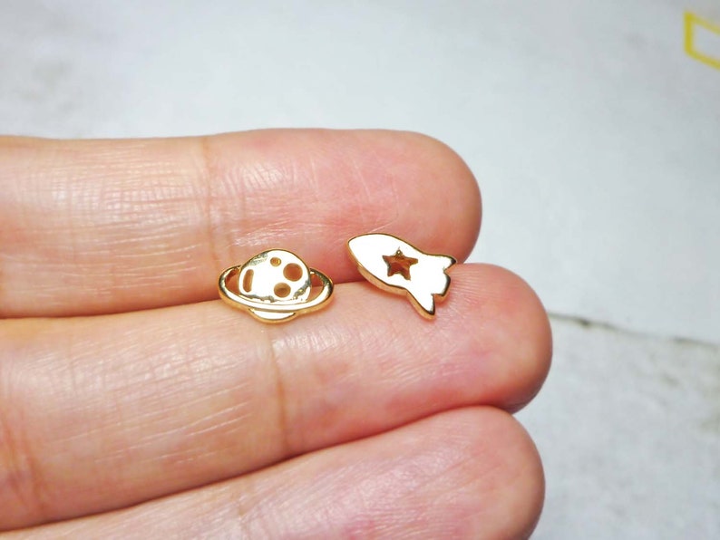 May include: A pair of gold-toned earrings featuring a rocket and a planet design. The rocket has a star cut-out and the planet has a ring around it.