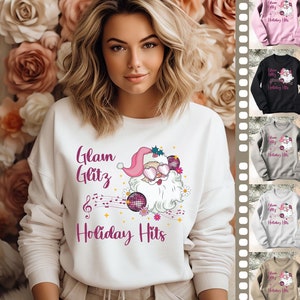 May include: A white sweatshirt featuring a Santa Claus graphic wearing sunglasses and a pink hat, with the text "Glam Glitz Holiday Hits" in pink. The design includes musical notes and disco balls. Additional sweatshirts in pink, black, gray, and tan are also shown.