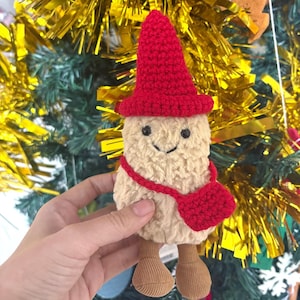 Puede incluir: Una muñeca de peluche de champiñón hecha a mano con un sombrero rojo y una bolsa a juego. El champiñón tiene una cara sonriente y pies de pana. La muñeca es beige y roja, y es una decoración festiva.