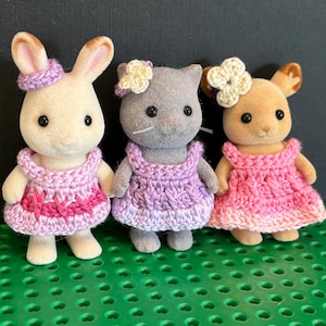 Puede incluir: Tres figuras de animales en miniatura: un conejito, un gato y un ciervo. Cada uno lleva un vestido de ganchillo en tonos rosas y morados, y un adorno floral a juego. El conejito es blanco, el gato es gris y el ciervo es marrón claro.