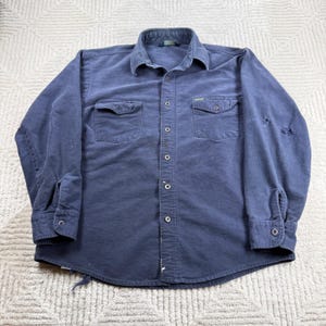Puede incluir: Una camisa de manga larga azul marino con botones, cuello clásico, dos bolsillos en el pecho con solapas abotonadas y puños con botones. La camisa muestra signos visibles de desgaste, incluyendo pequeños desgarros en la manga izquierda.