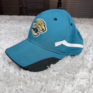 May include: Teal baseball cap with a team logo of a jaguar's head in yellow and black. The cap has a curved brim with a black underside, white stripes on the side, and black accents. The cap is made of a durable fabric.