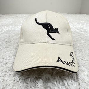 May include: Cream-colored baseball cap featuring a black kangaroo silhouette and the word "Australia" in a script font. The cap has a curved brim with a black trim and is made of a textured fabric.