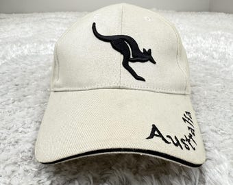 May include: Cream-colored baseball cap featuring a black kangaroo silhouette and the word "Australia" in a script font. The cap has a curved brim with a black trim and is made of a textured fabric.