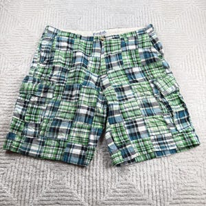 May include: Cargo shorts featuring a patchwork plaid pattern in shades of green, blue, white, and black. The shorts include a button closure, a zip fly, and two side cargo pockets with flaps. A white interior waistband label reads Plaid.