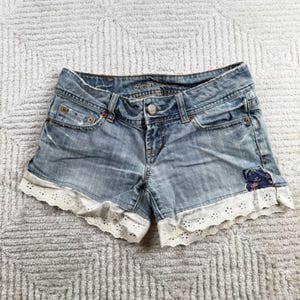 Puede incluir: Pantalones cortos de mezclilla azul claro con cintura baja, cierre de botón metálico y cinco bolsillos. Las aberturas de las piernas tienen un borde de encaje blanco con ojales y un pequeño parche de tela floral azul oscuro en la parte inferior.