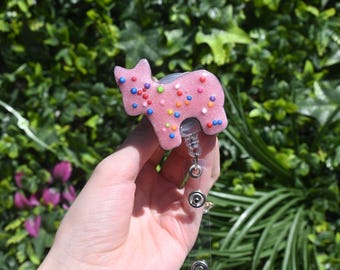 May include: Pink cookie-shaped badge holder with colorful sprinkles. The holder is attached to a retractable badge reel.