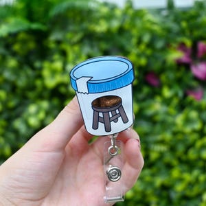 May include: A badge reel featuring a cartoon image of a stool inside a specimen cup with a blue lid. The badge reel has a clear plastic strap and a metal clip.