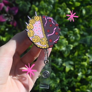 Może przedstawiać: Okrągła odznaka z podzielonym wzorem: jedna połowa to różowe ciasto z żółtym lukrem i posypką, druga połowa to czekoladowe ciasto z czerwonymi wiśniami i żelkowymi robakami. Rolka ma przezroczysty plastikowy klips i chowany sznurek.