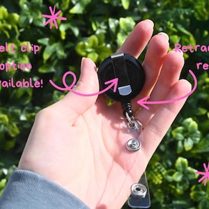 May include: A black retractable badge reel with a belt clip option. The reel is held in a hand against a backdrop of green foliage. The reel has a clear plastic strap with a snap closure.