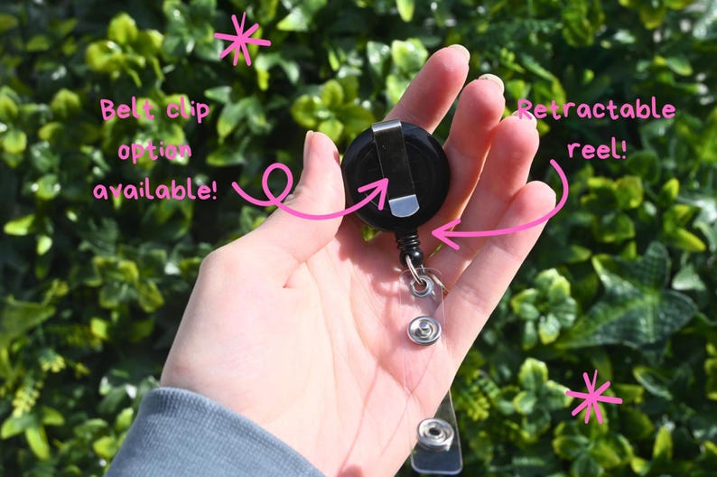 May include: A black retractable badge reel with a belt clip option. The reel is held in a hand against a backdrop of green foliage. The reel has a clear plastic strap with a snap closure.