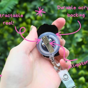 May include: A black badge reel with a retractable reel, durable acrylic backing, and an alligator clip. The reel is held in a hand against a blurred green background. Text on the image reads "Retractable reel!" and "Durable acrylic backing" and "alligator clip!"
