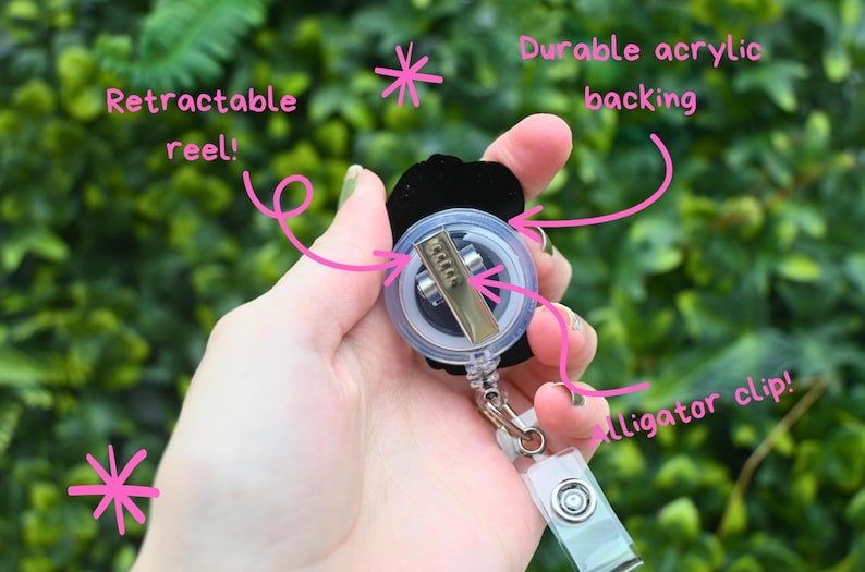 May include: A black badge reel with a retractable reel, durable acrylic backing, and an alligator clip. The reel is held in a hand against a blurred green background. Text on the image reads "Retractable reel!" and "Durable acrylic backing" and "alligator clip!"