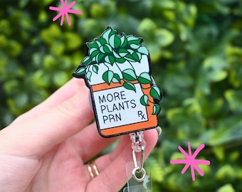 May include: A badge reel with a plant-themed design. The badge features a white and orange pill bottle shape with the words "MORE PLANTS PRN" and a green plant illustration. The reel has a clear plastic strap and a silver-coloured clip.