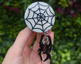 May include: A round, glittery badge reel with a black spiderweb design. A black spider charm with a red hourglass marking hangs below. The badge reel is silver and black, perfect for Halloween or spooky themed events.