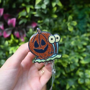 May include: A Halloween-themed badge reel featuring a smiling pumpkin with glitter accents and cartoon snail eyes. The pumpkin is orange with a black face, and the snail eyes are yellow and blue. The badge reel has a clear plastic strap.