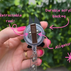 May include: A black and white badge reel with a retractable reel and alligator clip. The reel has a durable acrylic backing. The text on the image says "Retractable reel!" and "Durable acrylic backing" and "Alligator clip!"