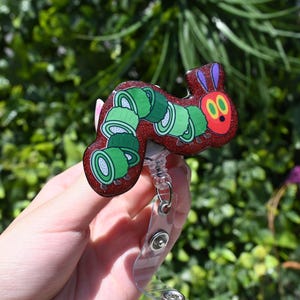 Peut inclure: Un porte-badge en paillettes rouge et vert représentant la Chenille qui fait très faim. La chenille est représentée avec une tête rouge et un corps vert, avec une série de cercles verts représentant la nourriture qu'elle a mangée.