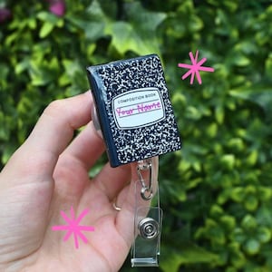 May include: A composition book-themed retractable badge holder. The badge reel features a miniature notebook design with a black and white speckled cover and the words "COMPOSITION BOOK" and "Your Name" on the front. The reel has a clear plastic strap and a metal clip.