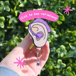 May include: A badge reel featuring a cartoon ear with a hearing aid. The ear is beige with purple accents, and a rainbow design. The text "Click to see variations!" is in a pink bubble.