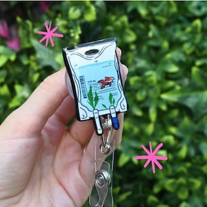 Può includere: Un porta badge con un design a cartone animato di sacca per flebo, che mostra un pesce rosso. La sacca ha dettagli blu e bianchi, un codice a barre e la scritta '150 ml'. Il porta badge è tenuto da una clip di plastica trasparente.
