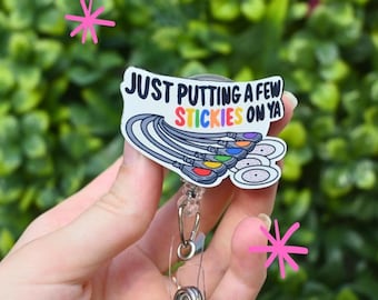 May include: A white badge reel with the text "JUST PUTTING A FEW STICKIES ON YA" and a design of colorful stickers. The reel has a clear plastic retractable cord and a metal clip.