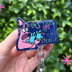 Op de afbeelding: Een badge reel met een stripfiguur en de tekst "DON'T TOUCH ME, I'M STERILE". De badge reel heeft een transparant intrekbaar koord van plastic en een metalen clip. De stripfiguur is roze en blauw.
