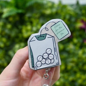 Puede incluir: Un portatarjetas con forma de bolsita de té, con un borde plateado brillante. El diseño de la bolsita de té contiene pastillas y el texto "Here for the pharma tea" en una pequeña etiqueta. El portatarjetas tiene un cordón retráctil de plástico transparente y un clip metálico.
