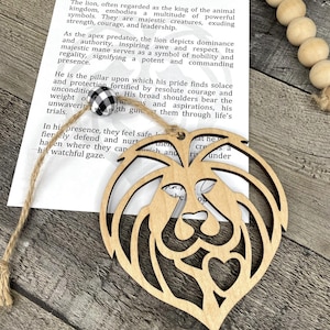 May include: Wooden lion-shaped ornament with intricate cut-out details, hanging from a twine string. The ornament is displayed with a printed card that reads "A Story of Strength & Protection". The background includes wooden beads.