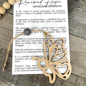 May include: A wooden butterfly-shaped ornament with a heart cutout, hanging on a string, rests on a card with the words "Renewed Hope" and text about resilience. The ornament is light brown.