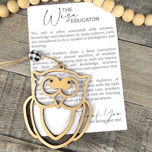 May include: A wooden owl ornament with the text "The Wise Educator" and a message about teachers. The owl is light brown with cut-out details. A black and white checkered bead is attached to the ornament.