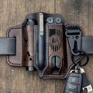 May include: A brown leather belt organizer with compartments for a flashlight, pen, and multi-tool. The organizer features a laser-etched American flag design and the name "ALEXANDER". A black belt and a key ring with keys and a knife are also visible.