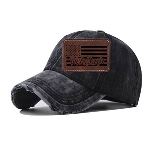 May include: Black baseball cap with a distressed finish. Features a brown leather patch with an American flag design and the text "We The People". The cap has a curved brim and an adjustable strap.