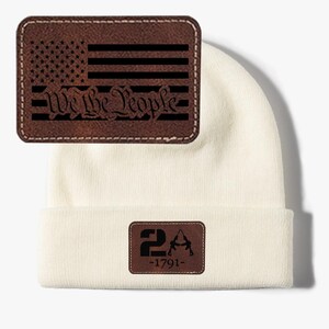 May include: Off-white beanie with two brown leather patches. One patch features a black American flag design with the text "We the People." The other patch displays "2A" and two rifles with the text "-1791-".