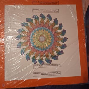 May include: A square white board with a vibrant, multi-colored geometric mandala pattern in the center, featuring shades of orange, blue, green, and yellow. The board is encased in a clear plastic bag with printed safety warnings in multiple languages.