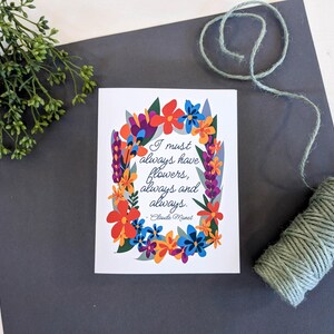 Può includere: Un biglietto bianco con una ghirlanda di fiori colorati e una citazione di Claude Monet che recita: "Devo sempre avere fiori, sempre e sempre."