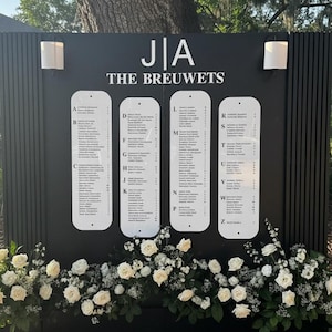 Peut inclure: Un plan de table de mariage noir avec des lettres blanches et quatre colonnes de noms, avec le texte "J|A THE BREUWETS" en haut. Des roses blanches et de la verdure décorent la base du plan.