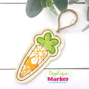May include: A fabric carrot-shaped ornament with a green leafy top and an orange and white patterned body. The carrot is outlined in black stitching and has a jute string for hanging. The Applique Market logo is visible.
