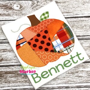 Op de afbeelding: Een wit canvas met een patchwork-pompoenontwerp en de naam "Bennett" geborduurd in groen. De pompoen is gemaakt van verschillende stoffen met verschillende patronen, waaronder ruit, stippen en geometrische vormen.