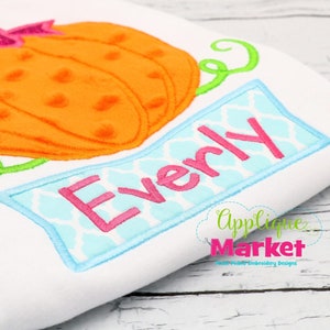 May include: Embroidered design featuring a pumpkin with a pink bow and a green stem, with the name "Everly" stitched on a blue banner with a white and blue pattern.