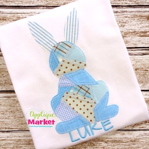 May include: A blue patchwork bunny applique with the name "LUKE" embroidered below. The bunny is made of different blue and brown fabrics with white polka dots and stripes.