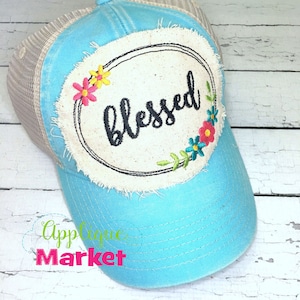 Puede incluir: Una gorra de camionero azul claro con un parche blanco que dice "blessed" en escritura negra. El parche está rodeado por un círculo negro y un diseño floral con bordados rosas, amarillos, azules y verdes.