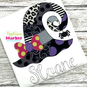Puede incluir: Una camiseta blanca con un sombrero de bruja de patchwork negro y morado con un lazo rosa y amarillo y una araña negra. El nombre "Sloane" está bordado en cursiva debajo del sombrero.