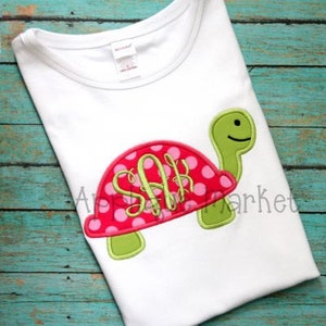 May include: A white t-shirt with a green and pink embroidered turtle with pink polka dots and the monogram "SAH" on the shell.