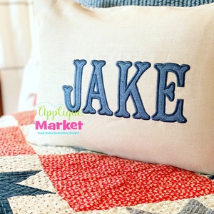 May include: A white cushion with the name "JAKE" embroidered in blue on a red, white and blue quilt. The cushion has the text "Applique Market" and "Fresh Picked Embroidery Designs" printed on it.