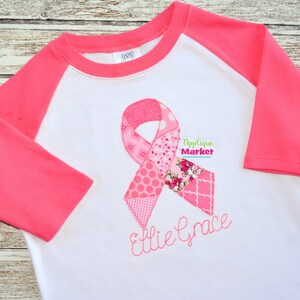 May include: A white and pink raglan-style shirt with a pink ribbon applique. The ribbon is made of various pink patterned fabrics and includes the name "Ellie Grace" embroidered in pink script. The shirt has pink sleeves.