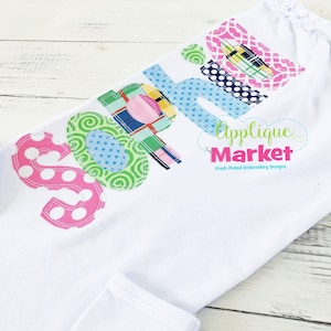Può includere: Una maglietta bianca con il nome "Sophia" scritto in lettere di stoffa colorate. Le lettere sono fatte di diversi motivi e colori, tra cui scozzese, pois e spirali. La maglietta ha anche il testo "Applique Market" e "Fresh Picked Embroidery Designs" stampato su di essa.