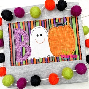 May include: A grey t-shirt with a colourful appliqué of a ghost, pumpkin, and the word "BOO" on a striped background. The shirt is decorated with a garland of colourful pom-poms.