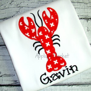 May include: White t-shirt featuring a red lobster design with white cross accents. The lobster has black antennae and legs. The name "Gavin" is embroidered in black script below the lobster. The text "Applique Market" is also visible.