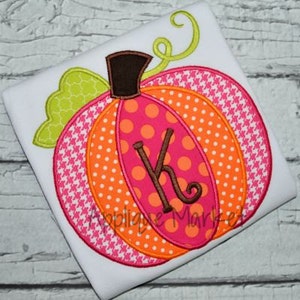 May include: A pink, orange, and white pumpkin with a brown stem and green leaves. The pumpkin has a pink and white polka dot pattern and a brown letter "K" in the center.